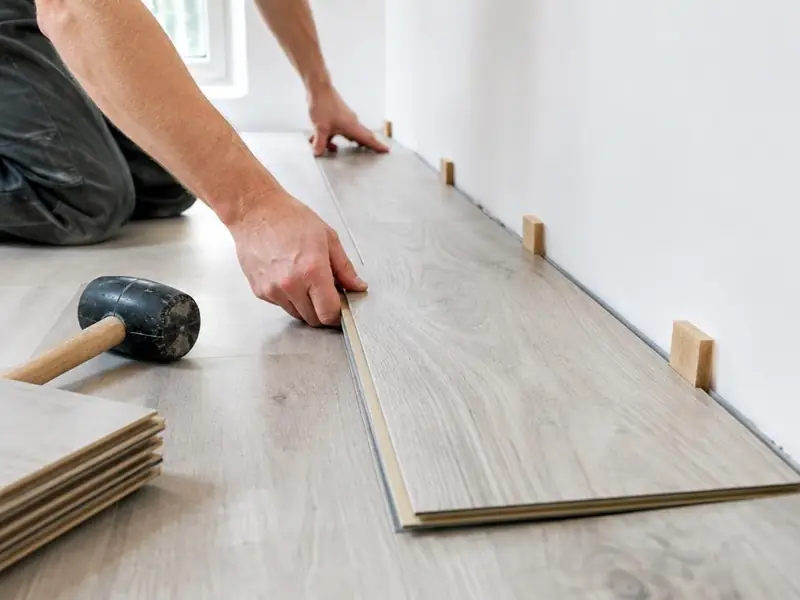 Mains posant la première rangée de lames de stratifié gris clair contre un mur avec des cales de dilatation.